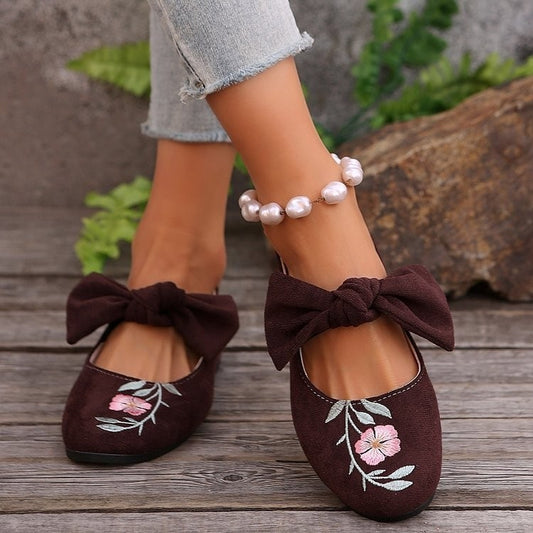 Brown shoes with floral embroidery and large bows worn on a wooden floor.