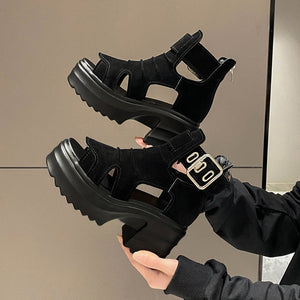 Black platform sandals with a gold buckle held by a person against a neutral background
