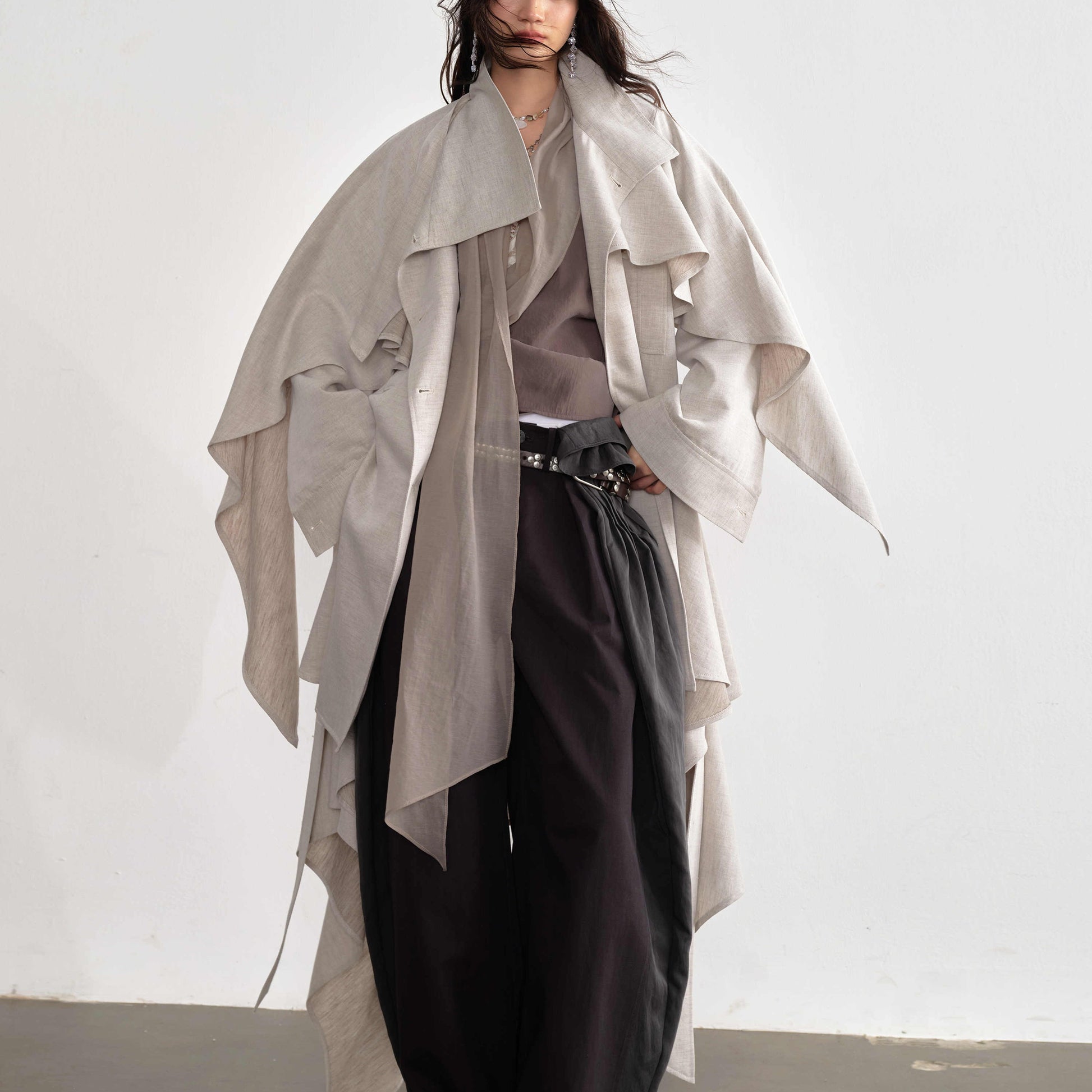 Person wearing a light gray draped coat over a neutral outfit on a white background