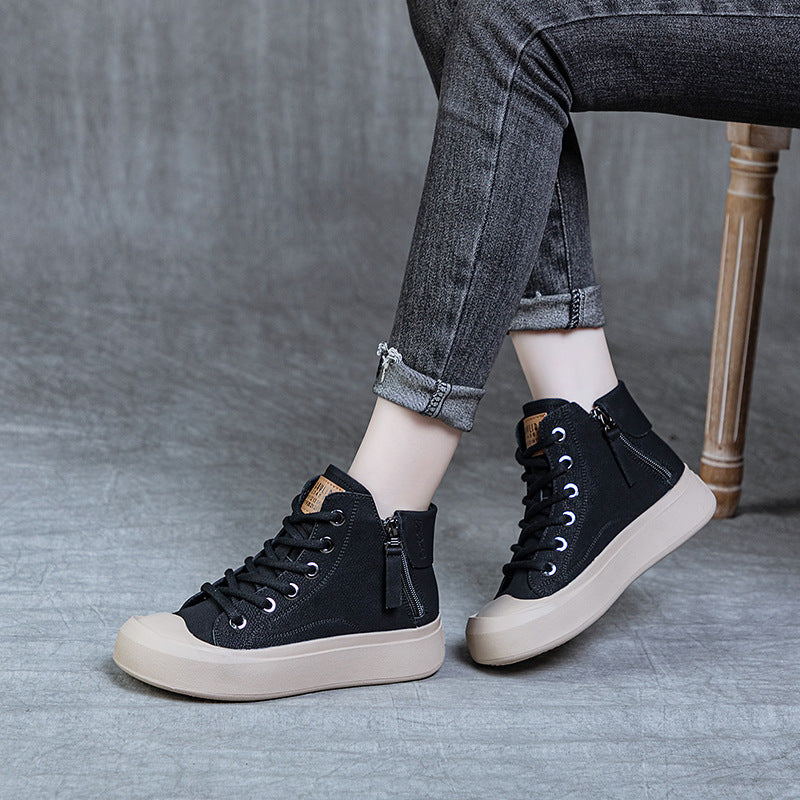 A person seated on a wooden chair wearing black high-top ankle boots with side zippers, lace-up fronts, and thick beige soles, paired with dark gray cuffed jeans against a gray studio backdrop.