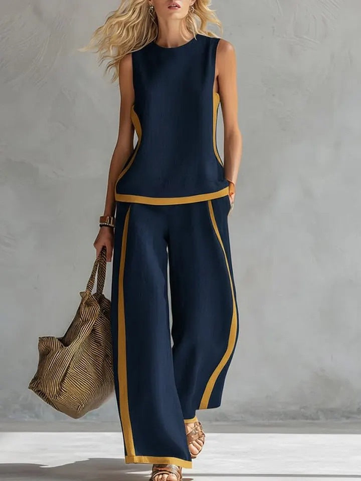 Woman wearing a navy sleeveless top and wide-leg pants with yellow accents, holding a woven bag against a gray background.