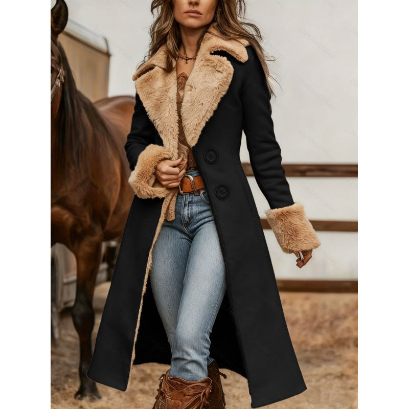 A woman models a long black coat with a thick beige faux-fur collar, cuffs, and lining, paired with blue jeans and brown boots in a barn setting with a horse in the background.