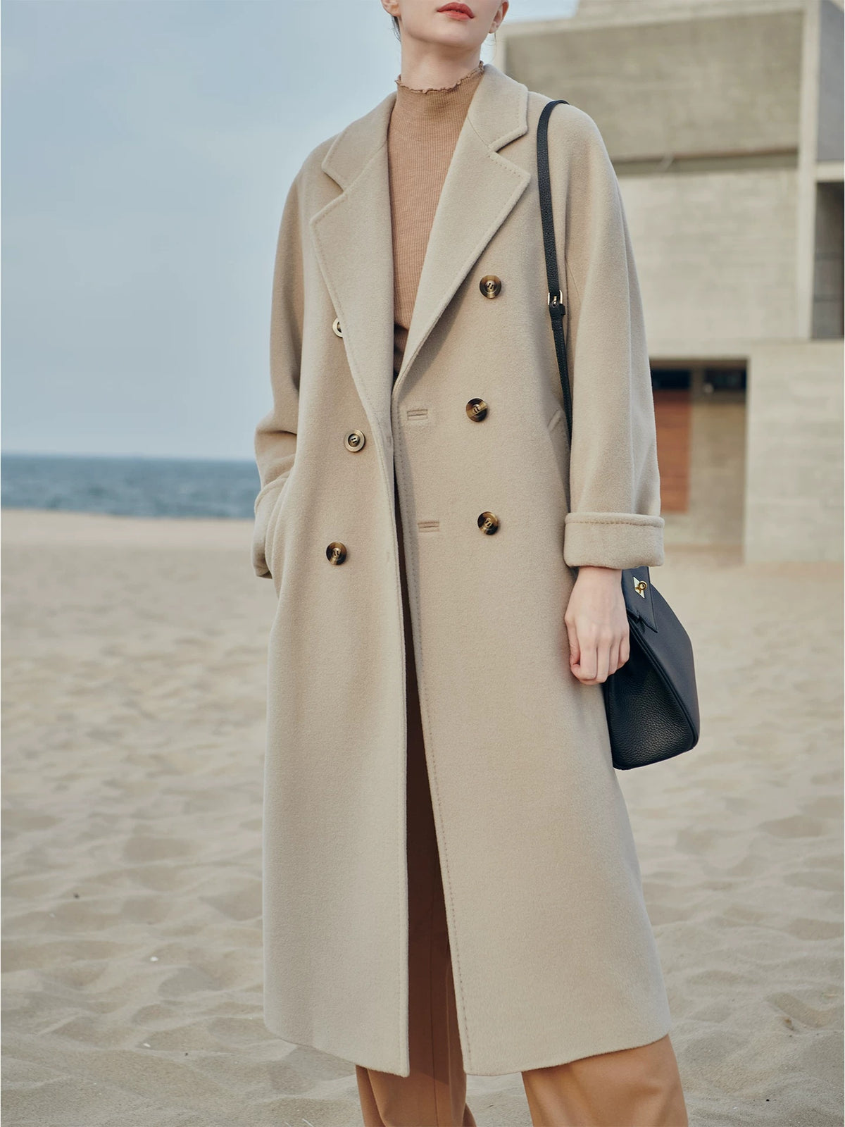 A woman standing on a sandy beach wearing a long double-breasted beige coat over a tan top, with a black shoulder bag.