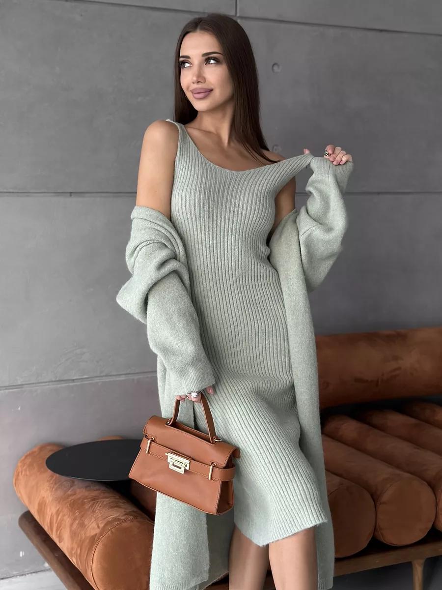 Woman in a light gray sweater dress holding a brown handbag against a gray wall.