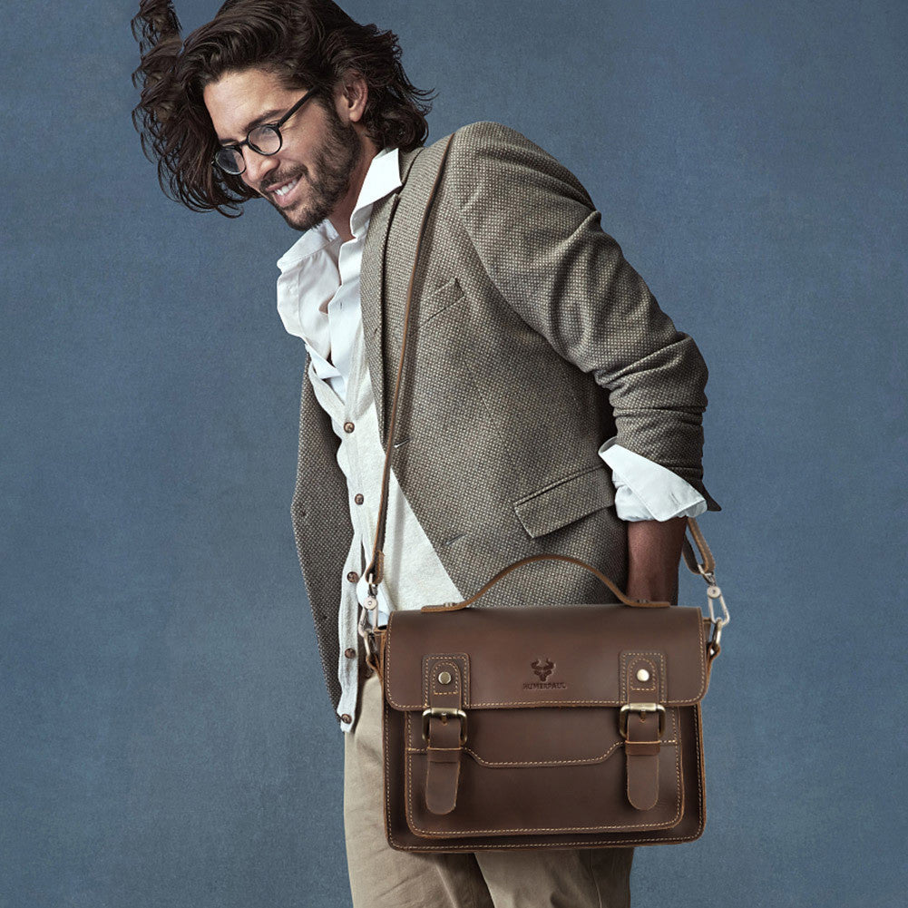 A man wearing a brown tweed blazer, white shirt, and beige trousers carries a brown leather satchel bag with a top handle and shoulder strap against a textured blue-gray background.