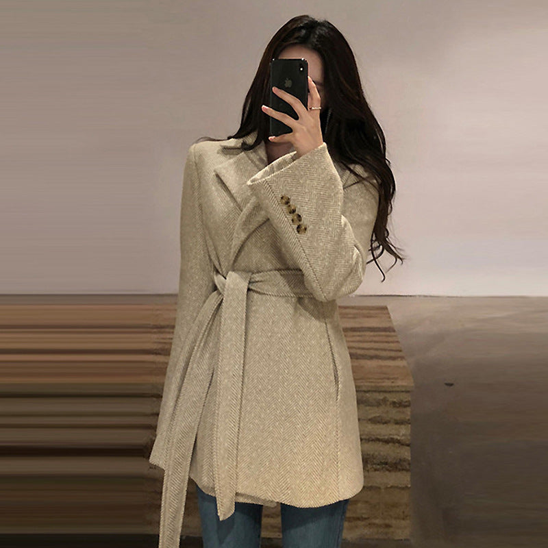 Woman wearing a beige coat taking a mirror selfie indoors.