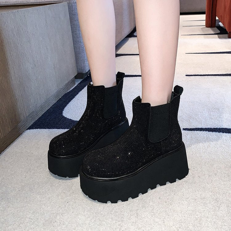 Black platform ankle boots covered in sparkling rhinestones, shown on a model's feet in an indoor setting with a patterned carpet.