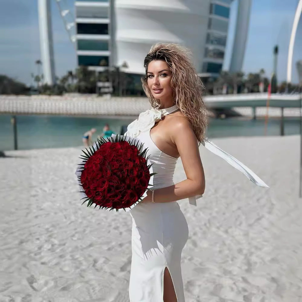 Woman in a white dress holding a large bouquet of red flowers on a beach with modern architecture in the background.