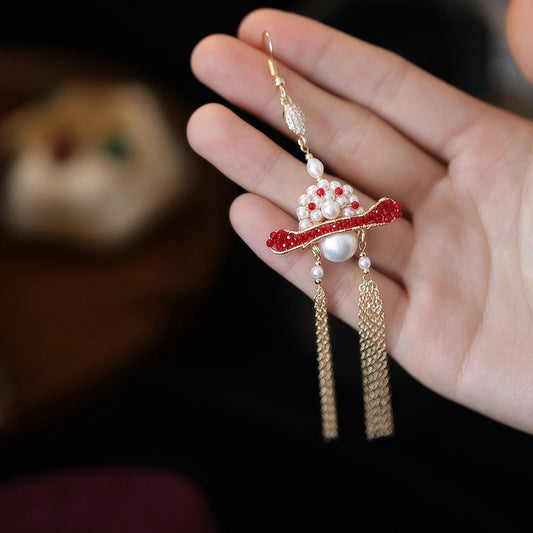 Decorative earring with red and gold design held by a hand against a blurred background