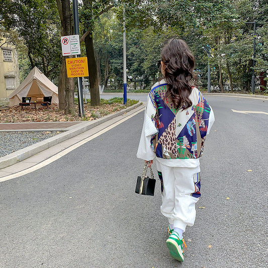 Person walking on a street wearing a colorful jacket and white pants.