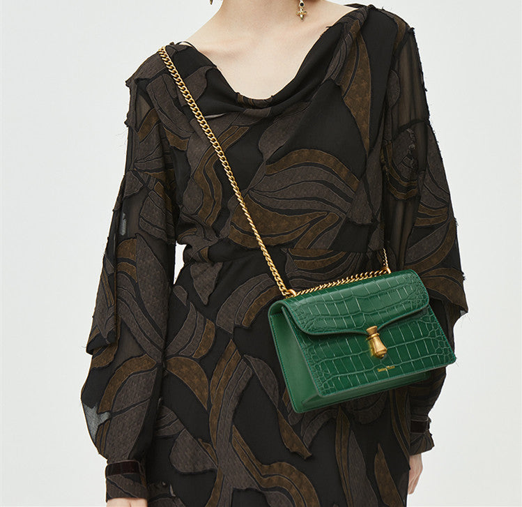 Green handbag with gold chain strap worn by a person in a black patterned outfit on a white background