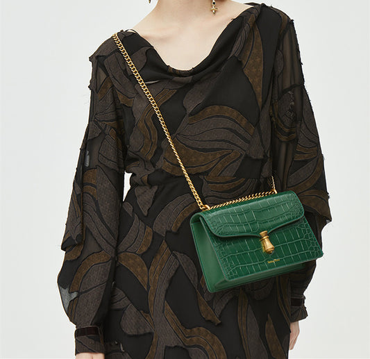 Green handbag with gold chain strap worn by a person in a black patterned outfit on a white background