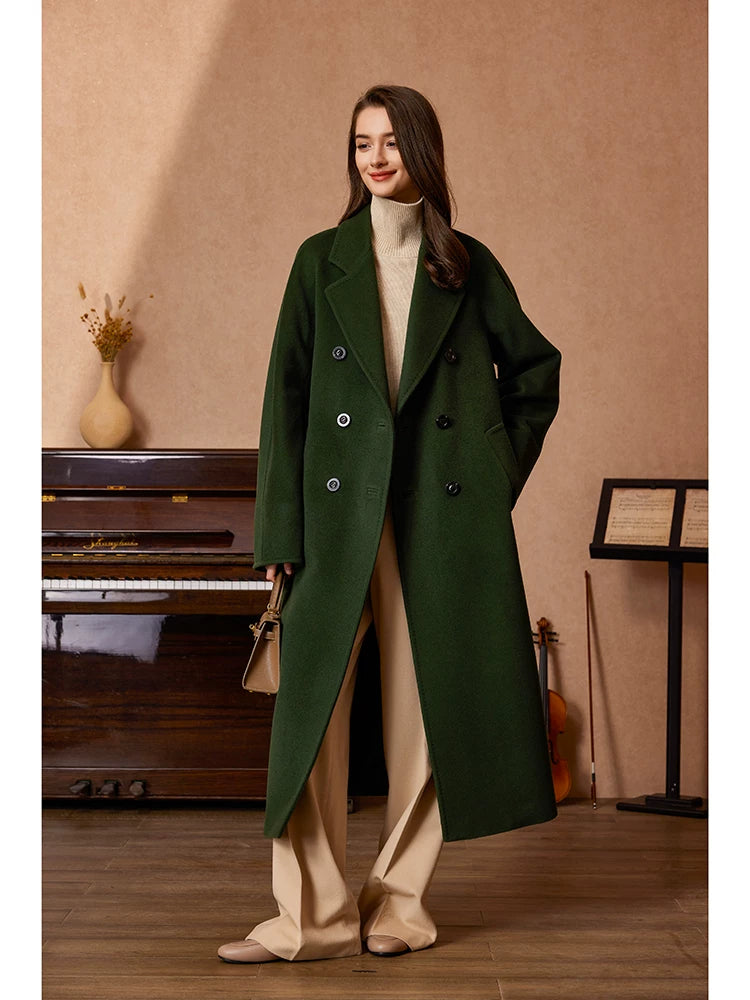 A woman stands in a warm indoor room wearing a long dark green double-breasted overcoat over a cream turtleneck and beige wide-leg pants. She holds a small beige handbag, with a piano, violin, and music stand in the background.