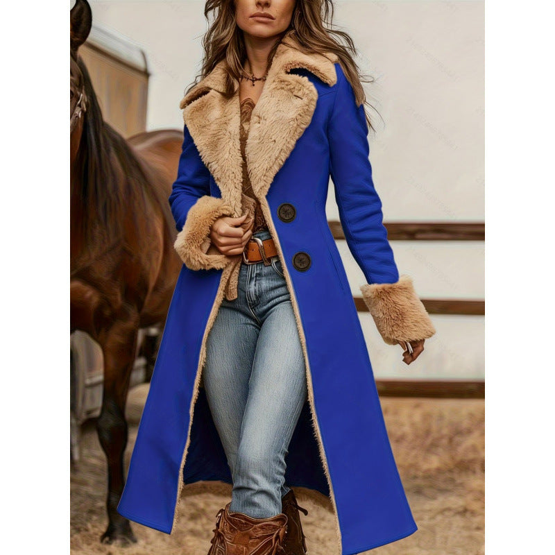 A woman wearing a long royal blue coat with a beige faux-fur collar and cuffs, paired with blue jeans and brown boots, standing in front of a horse in a rustic indoor setting.
