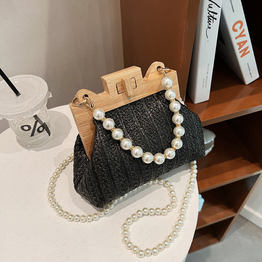 Black handbag with wooden handle and pearl chain on a surface with books and a cup in the background.