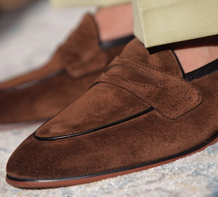 Brown suede loafers worn with beige pants on a textured surface