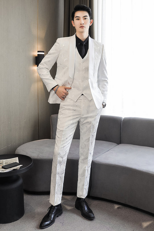 Man in a white suit standing in a modern room with a gray sofa and lamp.
