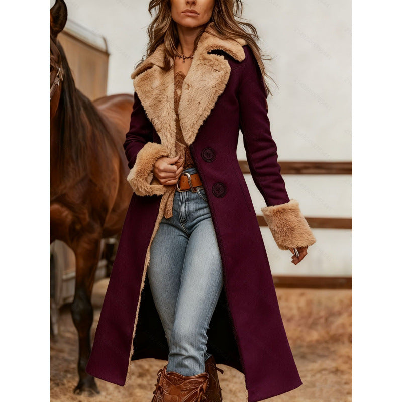 A woman wearing a long burgundy wool-blend coat with a beige shearling-style collar, cuffs, and lining, paired with light blue jeans and brown boots, standing in a barn with a horse in the background.