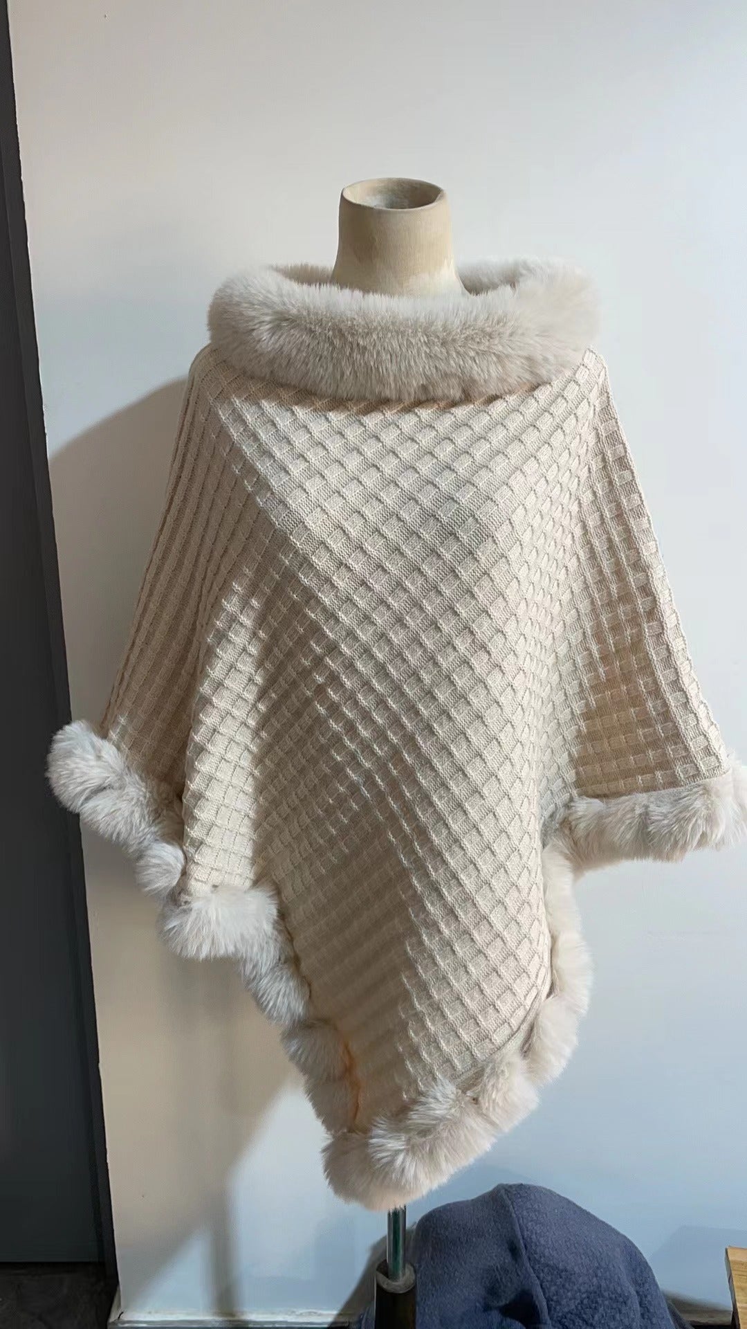 Knitted poncho with pom-poms on a mannequin against a white wall