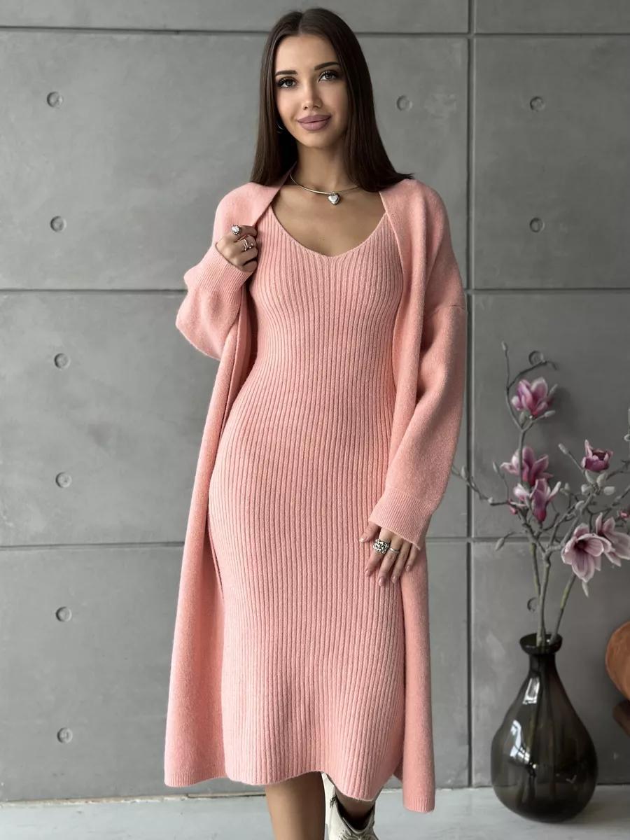 Woman wearing a pink dress and cardigan against a gray wall with a vase of flowers.