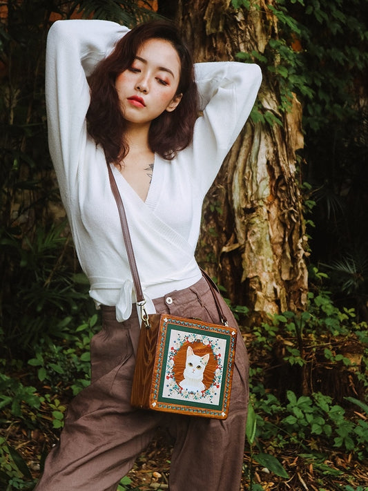 Woman holding a white cat in her arms with a handbag featuring a cat design in a forest setting