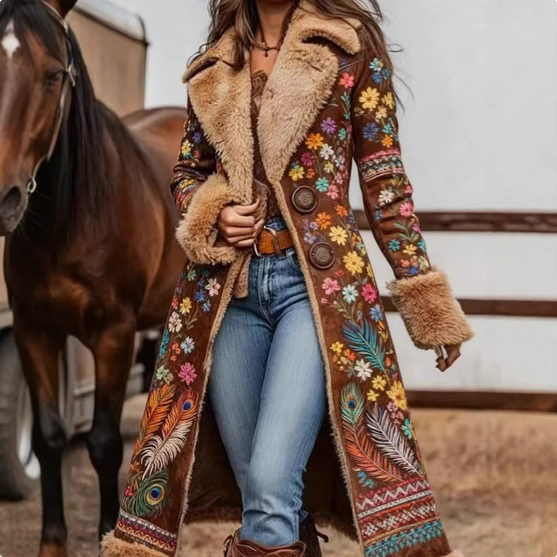 A woman wearing a long brown suede-like coat with colorful floral and feather embroidery, faux-fur trim, and large buttons, standing in a barn next to a brown horse.