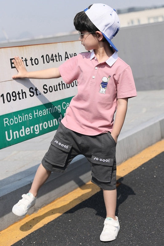 Child wearing a pink polo shirt with a cartoon character and gray shorts, standing on a street.