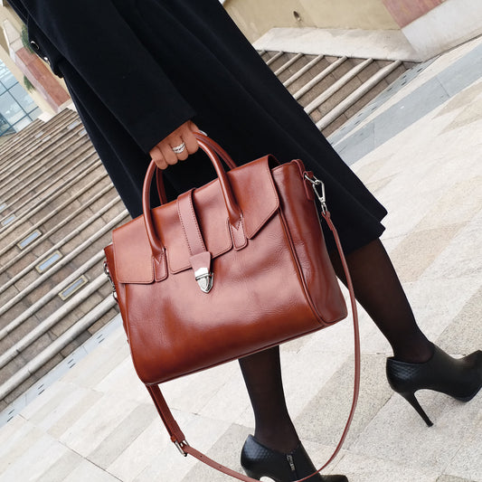 Brown leather handbag held by a person wearing a black coat and high heels on a city street.