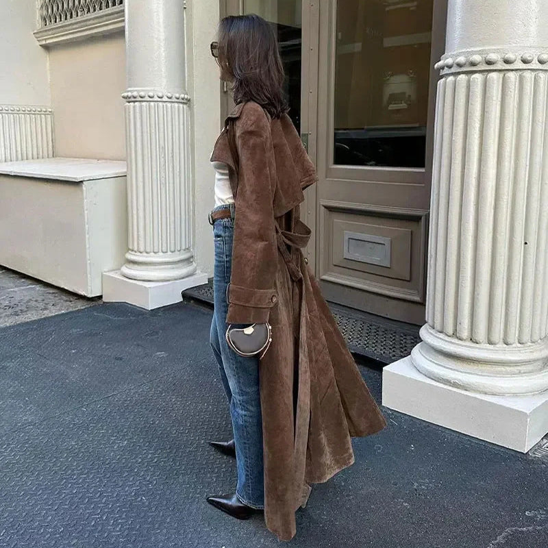 Person wearing a brown coat and blue jeans standing in front of a building entrance. By Millivan 