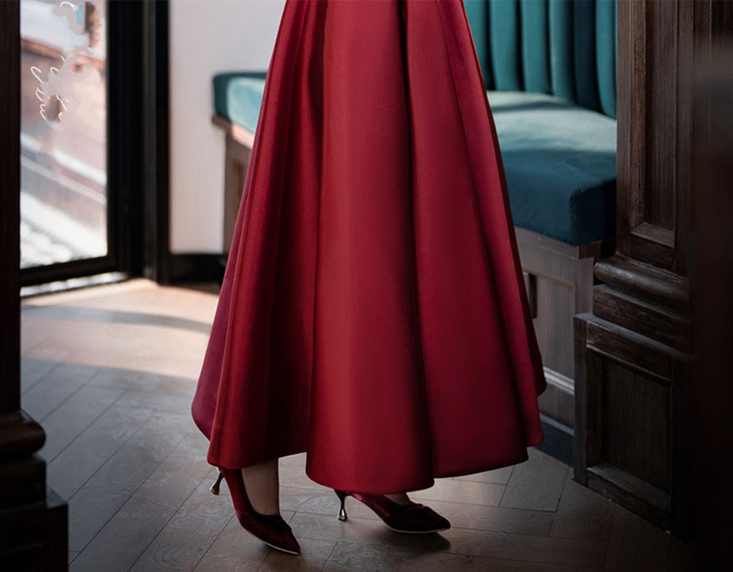 Person wearing a long red dress standing in a room with wooden floor and teal cushioned bench.