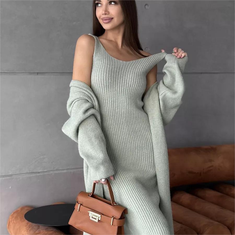 Woman wearing a long light gray sweater over a dress, holding a brown handbag against a gray wall.