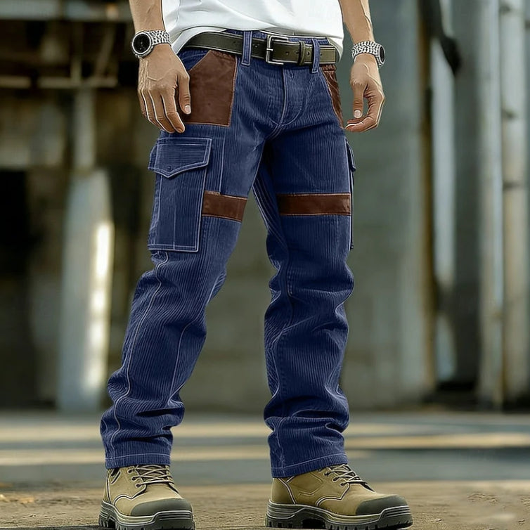 A person wearing blue corduroy cargo work pants with brown leather patch panels, styled with a white t-shirt, black belt, and tan work boots in an industrial setting.