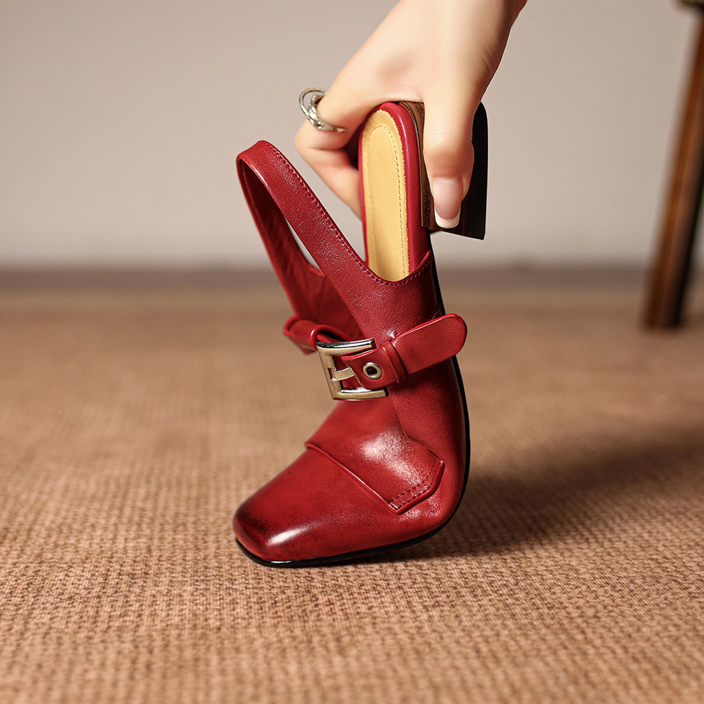 Red high-heeled shoe with a gold buckle held by a hand on a textured surface.