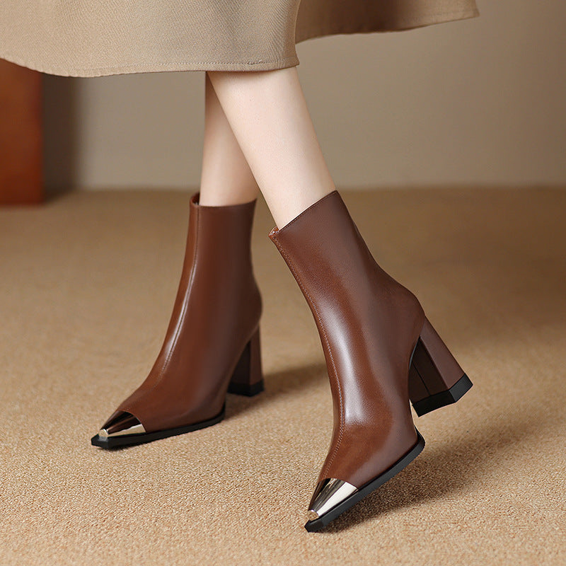 A pair of brown ankle boots with a pointed toe and metallic gold toe caps, shown on a model wearing a beige skirt against a neutral beige background.