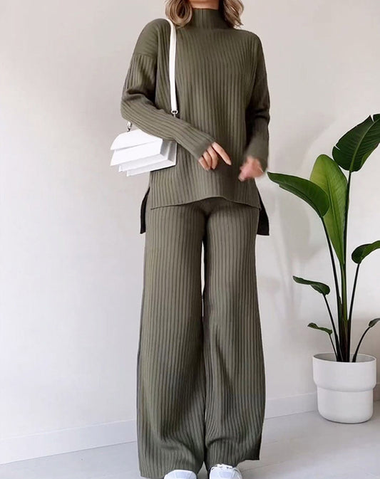Person wearing a green ribbed sweater and pants with a white bag, standing next to a potted plant on a white background.