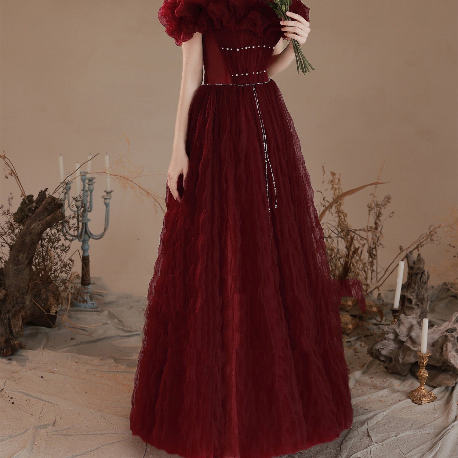 Woman in a burgundy evening gown holding flowers in a decorative setting with candles and plants.