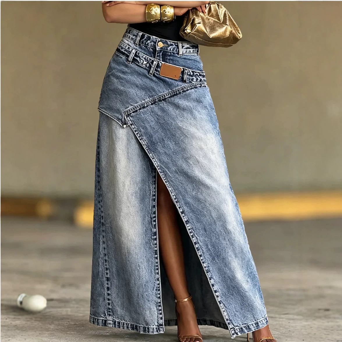 A woman models a long light-wash denim maxi skirt with an asymmetrical wrap-style front slit and a small brown leather accent patch near the waistband. She is wearing a black top, gold bracelets, and holding a metallic gold clutch against a neutral concrete wall background.
