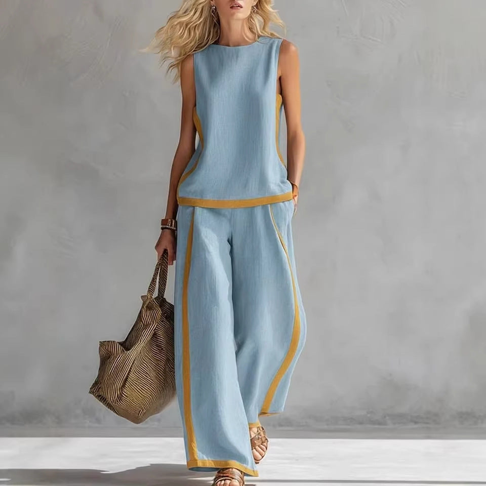Woman wearing a light blue sleeveless top and wide-leg pants with yellow trim, holding a woven bag against a gray background.