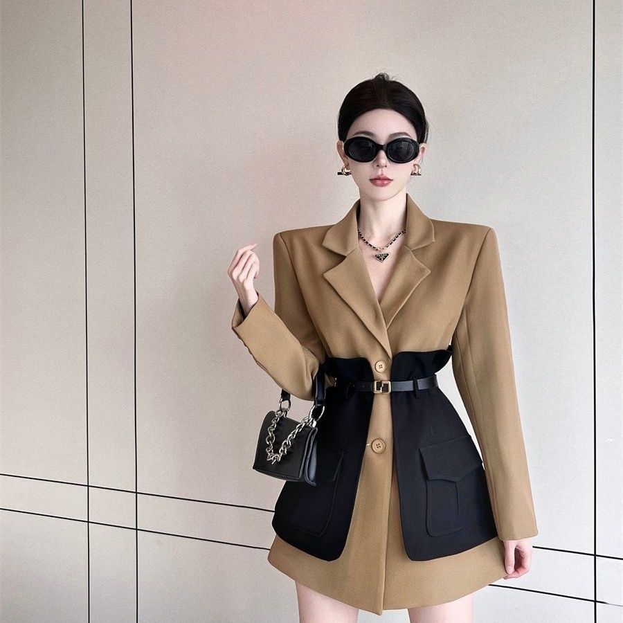 Woman wearing a beige coat over a black dress with sunglasses and a handbag against a light background