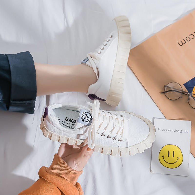 White sneakers with beige soles held by a person, with a white background and a box and glasses in the background.