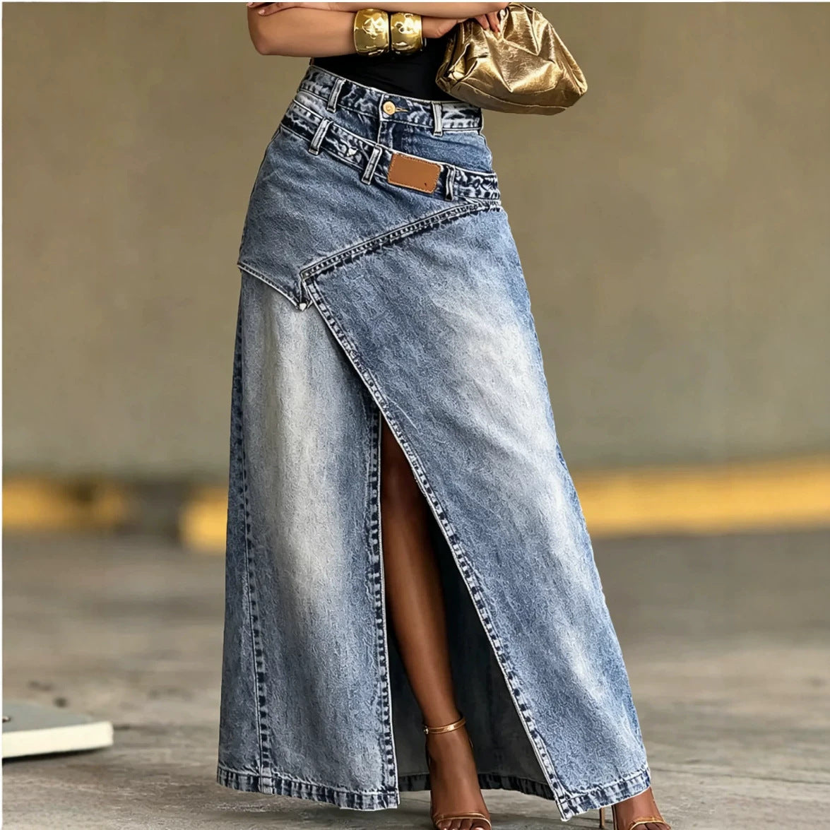 A woman wearing a long light-wash denim skirt with a high front slit, styled with a black top, gold bracelets, a metallic gold clutch, and brown heeled sandals.