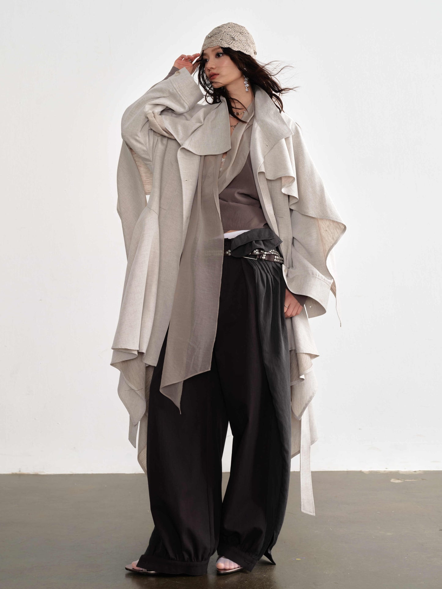Woman wearing a long, light-colored coat over a gray top and black pants on a white background