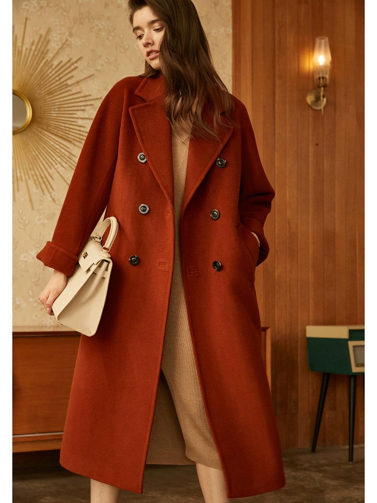 A woman wearing a long double-breasted rust-orange wool coat over a beige dress, holding a cream-colored handbag in a warm, wood-paneled interior setting.